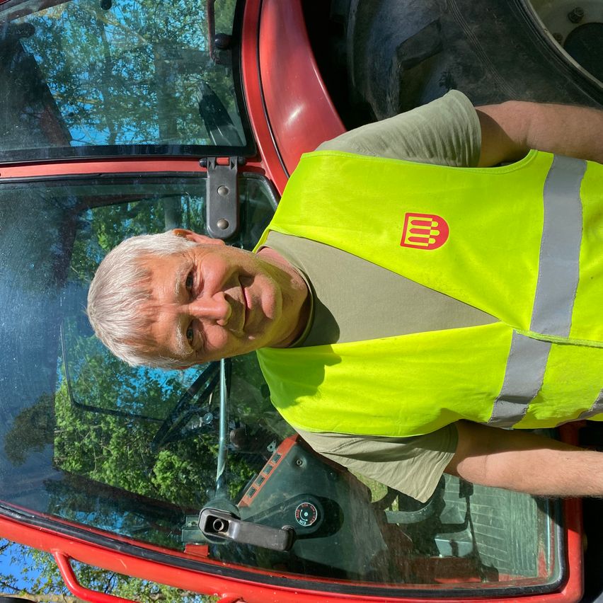Ein älterer Mann in einer neonfarbenen Sicherheitsweste lächelt, während er sich an einen roten Traktor lehnt, mit einem großen Reifen im Hintergrund und einem sonnigen, blauen Himmel.