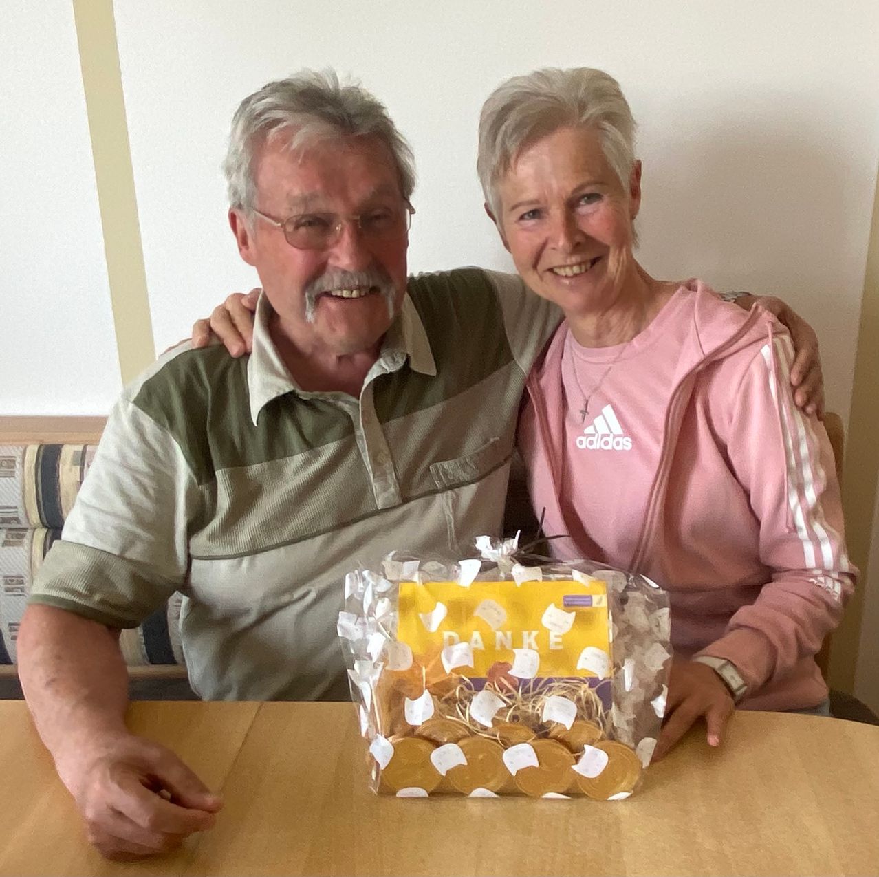 Ein älteres Paar lächelt, während es mit einer Tüte goldener Münzen an einem Tisch sitzt, einer hält eine Geschenktüte mit dem Wort Danke.