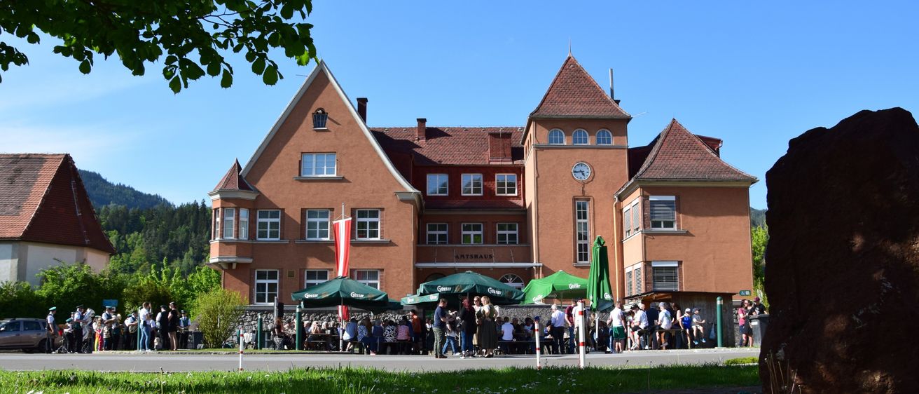 Ein großes braunes Gebäude mit einer Uhr, umgeben von Menschen mit grünen Schirmen und einem Rasen mit weißen Blumen.