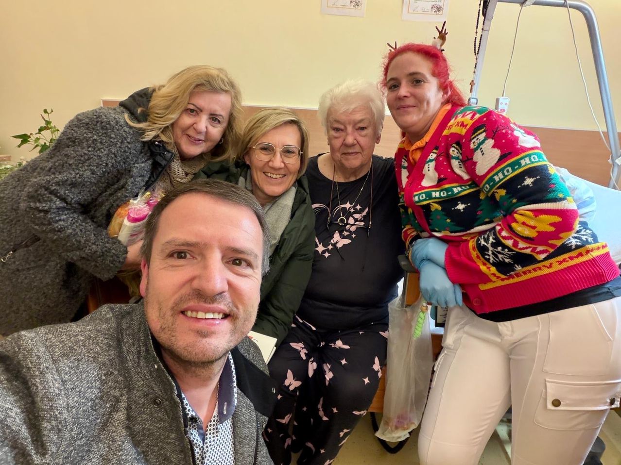 Fünf Erwachsene in einem Raum mit einer Frau im Krankenhausbett, eine Frau mit Rentierhörnern, ein Mann lächelt und drei Frauen lächeln.