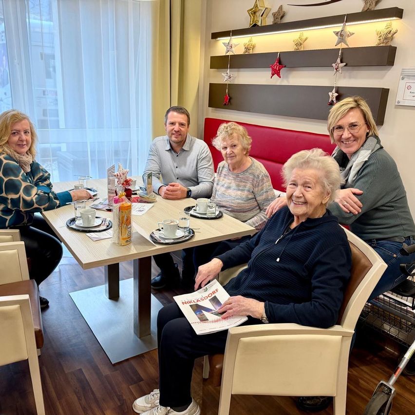 Eine Gruppe von Erwachsenen sitzt um einen Tisch in einem Restaurant. Sie haben Tassen, Teller und eine Zeitschrift. Eine Frau mit Brille sitzt mit ihren Armen um eine ältere Frau.
