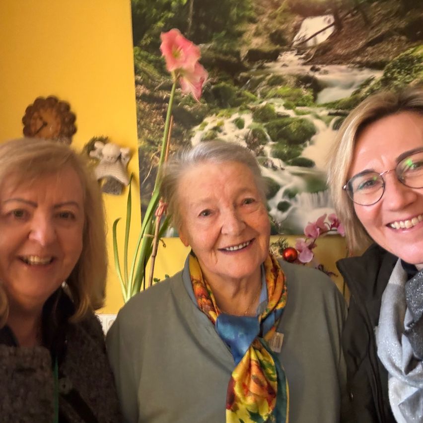 Drei Frauen lächeln und posieren für ein Foto mit einem malerischen Wasserfall im Hintergrund. Die Frau in der Mitte trägt ein farbenfrohes Tuch.