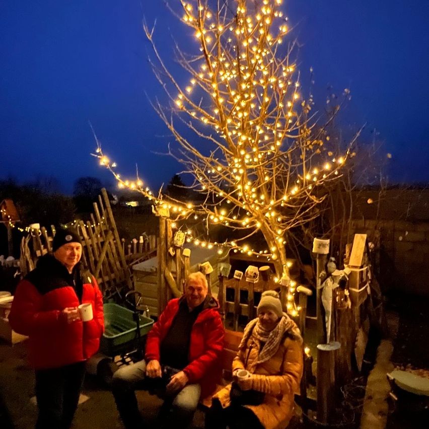 Drei Personen sitzen unter einem mit Lichtern geschmückten Baum. Zwei sitzen auf einer Bank und lächeln, während die dritte einen Becher in der Hand hält. Im Hintergrund befindet sich ein Holzzaun und ein schwach beleuchteter Himmel.