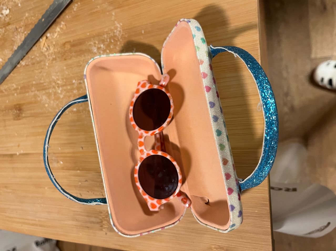 Eine rosa und weiße Sonnenbrillenetui mit glitzernden Griffen und Herzmuster ist geöffnet und zeigt ein Paar orange und schwarze Sonnenbrille.