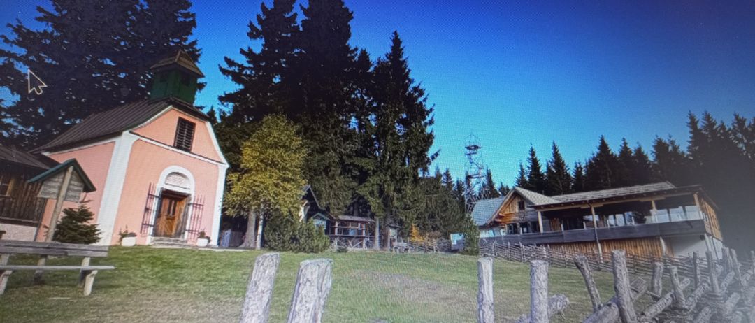 Eine ländliche Landschaft mit einem Holzzaun, einem Haus mit Turm und einer Blockhütte mit Veranda, umgeben von Bäumen und einem klaren blauen Himmel.