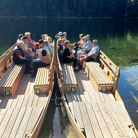 Eine Gruppe von Menschen genießt eine Fahrt auf einem hölzernen Boot auf einem ruhigen See mit einem bewaldeten Hintergrund.