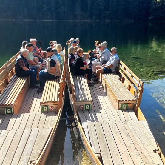 Eine Gruppe von Menschen genießt eine Fahrt auf einem hölzernen Boot auf einem ruhigen See mit einem bewaldeten Hintergrund.