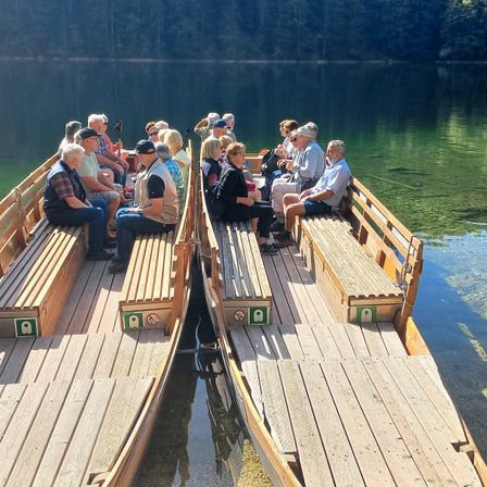 Eine Gruppe von Menschen sitzt auf Bänken auf einem Boot im See. Sie tragen alle Hüte und einige halten Weingläser. Das Boot ist aus Holz und hat ein Schild an der Seite.