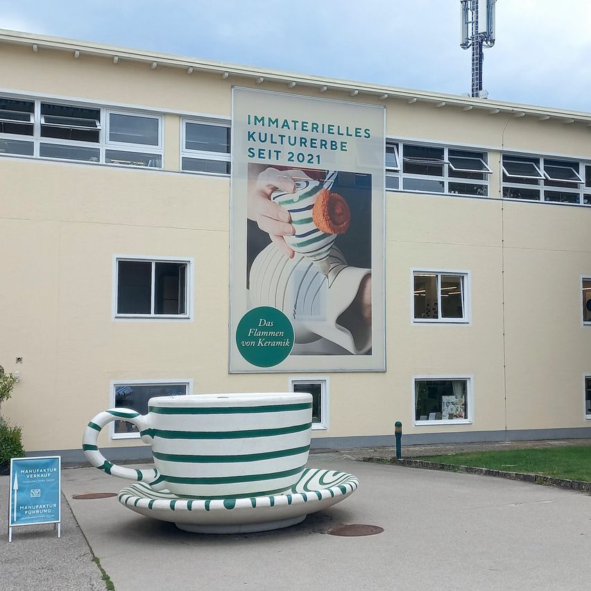 Eine große Keramiktasse und Untertasse stehen vor einem Gebäude mit einem Banner, das 'Immaterielles Kulturerbe seit 2021' besagt. Das Banner zeigt eine Hand, die ein Keramikobjekt hält.