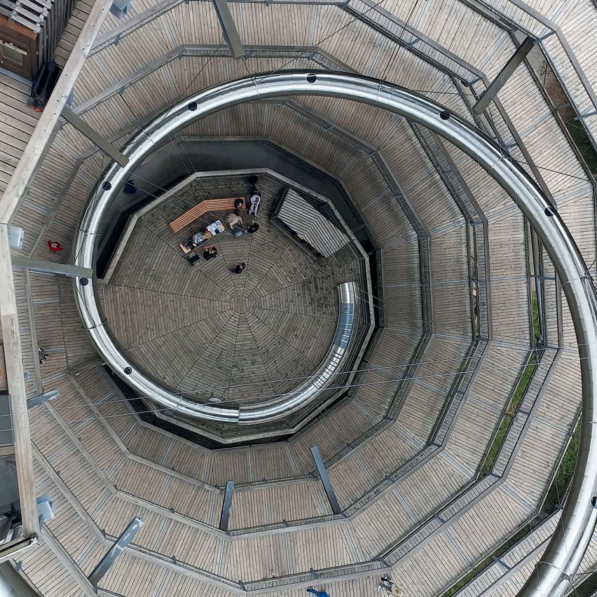 Ein Blick von oben auf eine spiralförmige Struktur mit Menschen, die auf Bänken um einen zentralen Bereich herum sitzen. Die Struktur hat Holzböden und Metallgeländer.