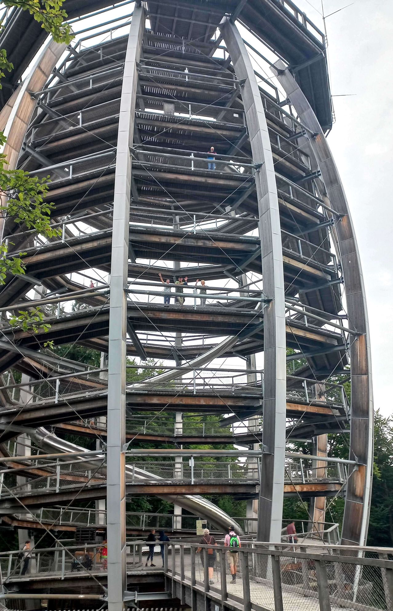 Eine spiralförmige Treppe aus Metall und Holz, mit Menschen auf verschiedenen Ebenen. Die Struktur ist von Bäumen umgeben.