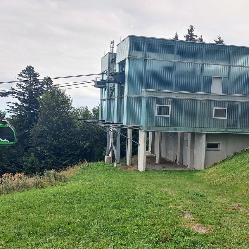 Ein modernes Gebäude mit Glaswänden und einer daran befestigten Seilbahn, auf einem grünen Hügel mit Bäumen im Hintergrund.