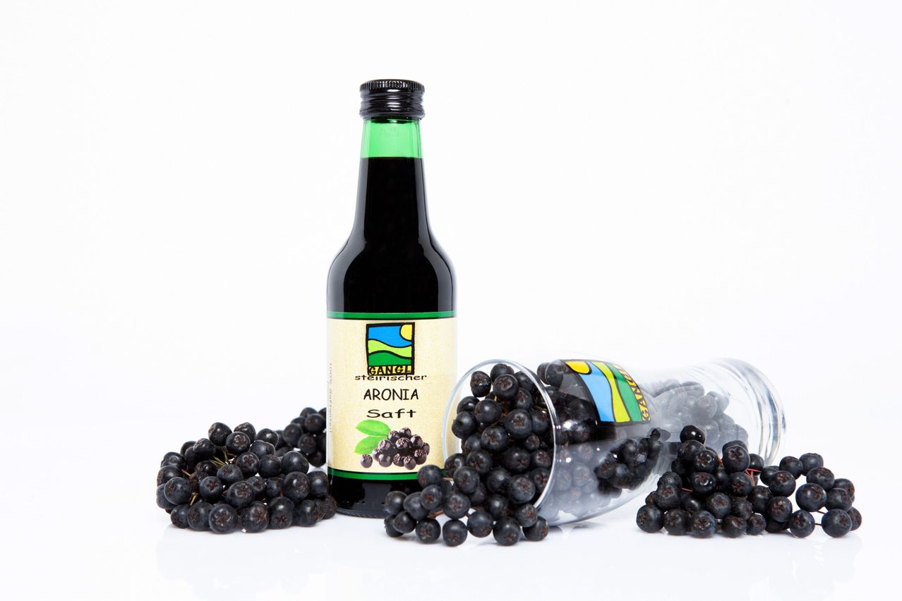 Eine Flasche Aroniasaft mit frischen schwarzen Beeren in einem klaren Plastikbehälter und einem kleinen Haufen Beeren.