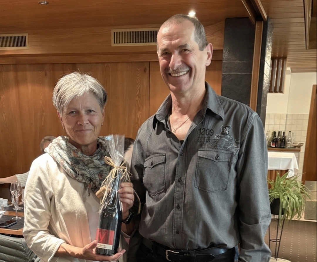 Ein Mann und eine Frau stehen lächelnd in einem Raum. Die Frau hält eine Flasche Wein in Plastik verpackt. Hinter ihnen ist eine Holzwand und ein Luftventil zu sehen.