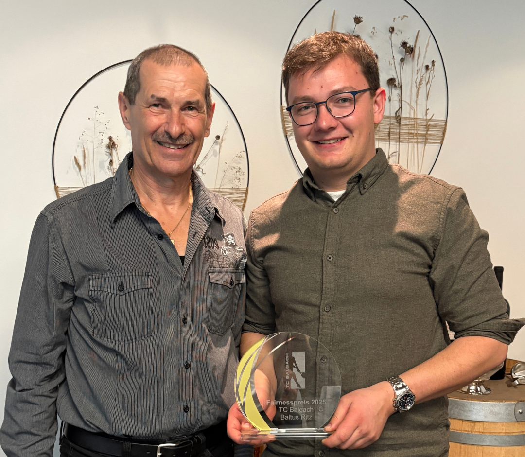 Zwei Männer stehen zusammen und halten einen Pokal. Der Mann auf der linken Seite hat ein Lächeln und trägt eine Halskette. Der Mann auf der rechten Seite trägt eine Brille und eine Uhr.