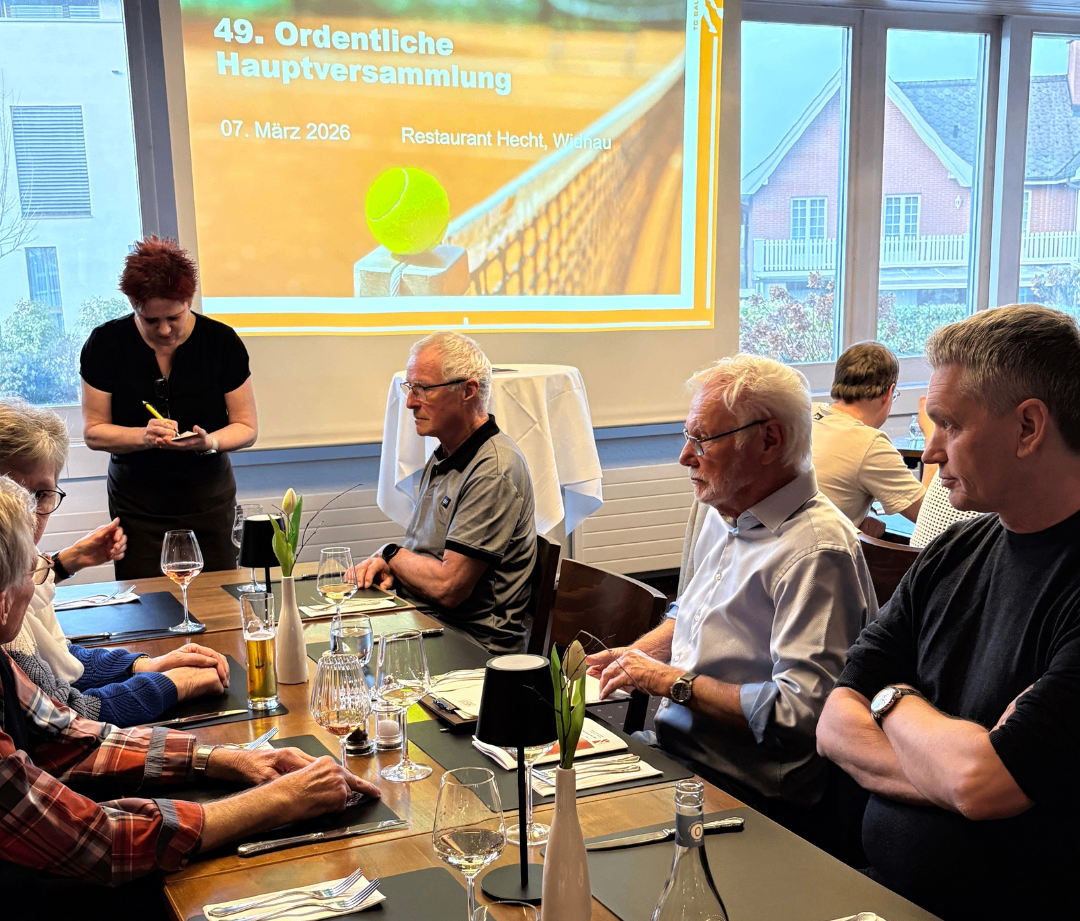 Eine Gruppe von Menschen sitzt an einem Tisch, möglicherweise in einem Restaurant. Eine Frau steht hinter ihnen und schreibt auf einem Notizblock. Eine Projektionsleinwand zeigt die Worte '49. Ordentliche Hauptversammlung 07. März 2026'.