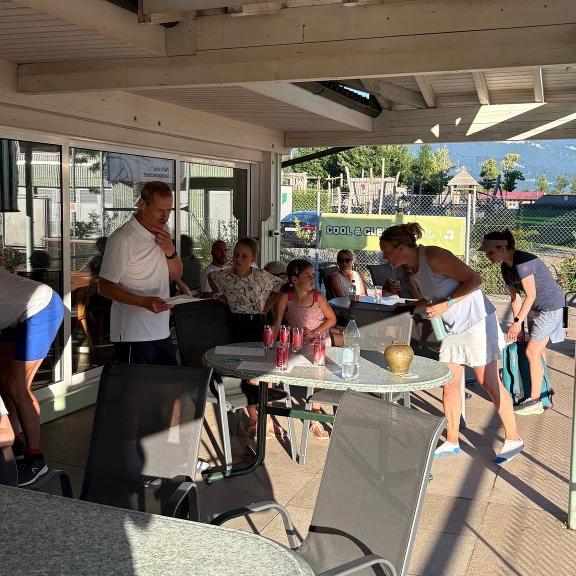 A group of people are gathered around a table in a shaded outdoor area. A man stands and speaks to them while others listen attentively. The table is set with drinks and papers. Behind them, a sign reads 'COOL & CLE'.