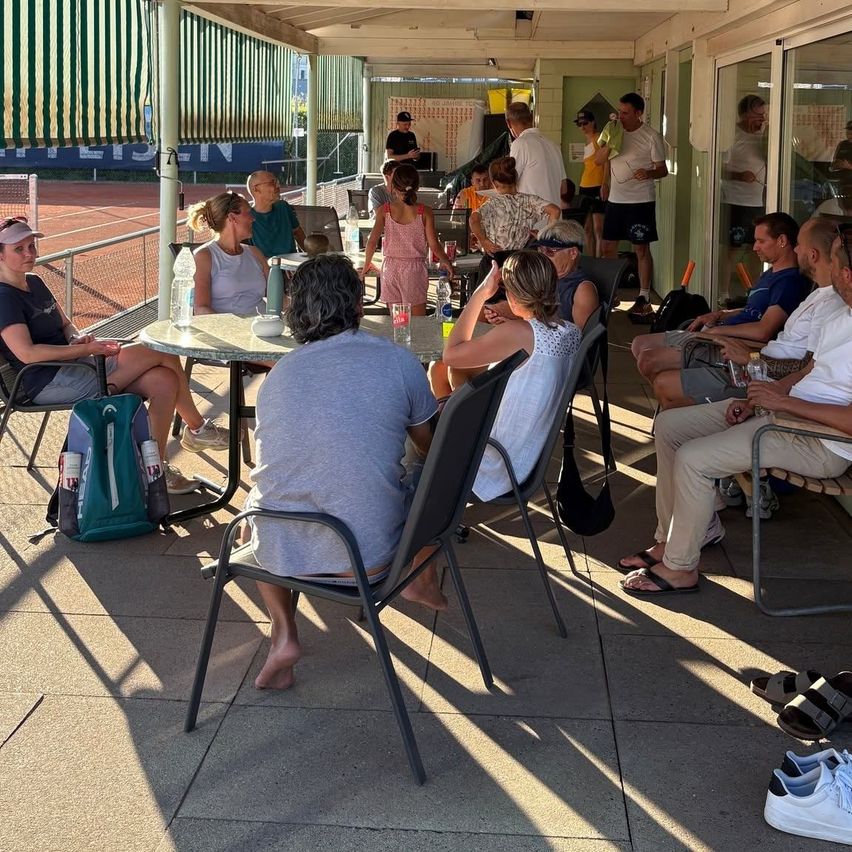 A group of people are sitting around a table in a shaded outdoor area, possibly at a tennis court. Some are wearing hats and glasses, and there are bottles and bags on the table.