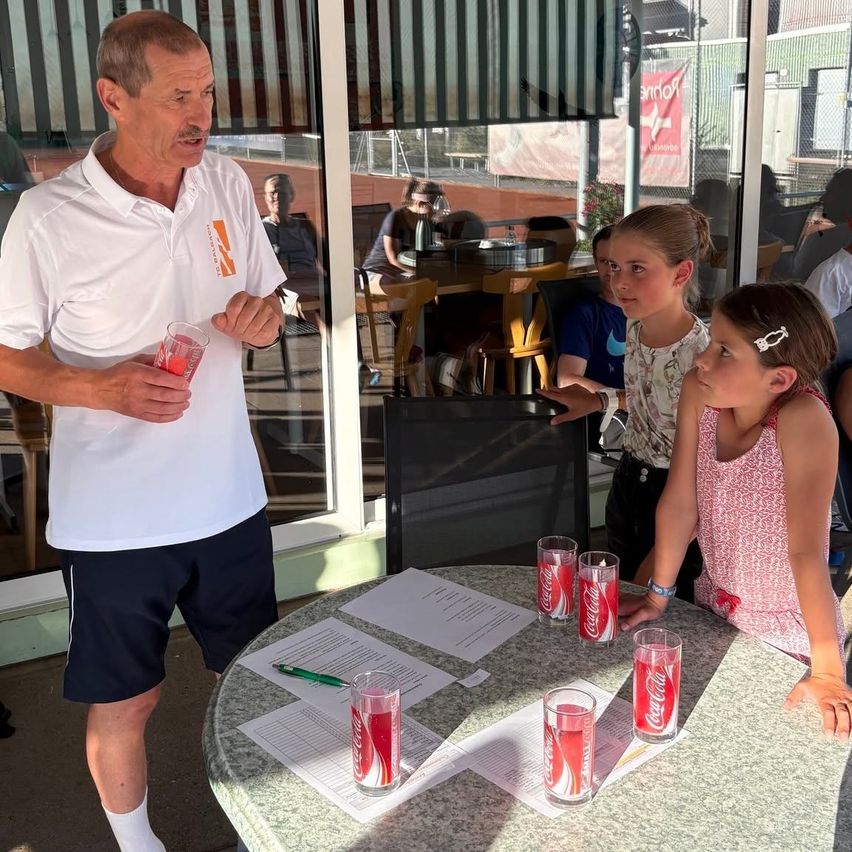 An older man in a white polo shirt stands at a table with four Coca-Cola glasses, speaking to two young girls. Papers and a pen are on the table. Behind them, a glass wall reflects a restaurant interior.