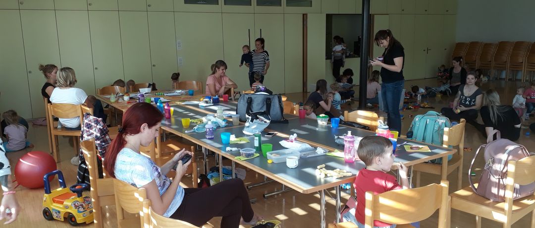 Eine Gruppe von Menschen, einschließlich Kindern, sitzt um Tische in einem Raum mit Holzböden herum. Einige essen, während andere auf ihre Handys schauen.