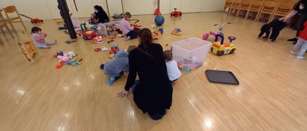 In einem Raum mit Holzboden spielen mehrere Kinder mit Spielzeug. Eine Frau kniet und interagiert mit den Kindern. Im Hintergrund befinden sich Schränke und ein Fenster.