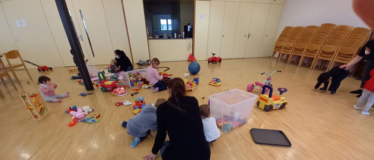 Eine Gruppe von Kindern und Erwachsenen befindet sich in einem Spielzimmer mit verstreuten Spielzeug. Eine Frau kniet und beobachtet ein spielendes Kind.