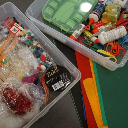 Zwei Plastikbehälter voller verschiedener Bastelutensilien, einschließlich Perlen, Bänder, Kleber und Scheren, auf einer grauen Oberfläche.
