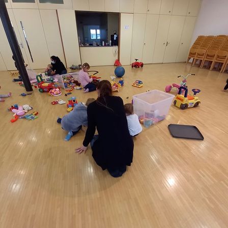 Mehrere Kinder spielen mit Spielzeug auf einem Holzboden. Eine Frau in einem schwarzen Kleid kniet und beobachtet die Kinder. In der Nähe gibt es mehr Spielzeug, darunter eine blaue Kugel.
