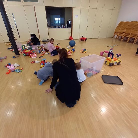 Mehrere Kinder spielen mit Spielzeug auf einem Holzboden. Eine Frau in einem schwarzen Kleid kniet und beobachtet die Kinder. In der Nähe gibt es mehr Spielzeug, darunter eine blaue Kugel.