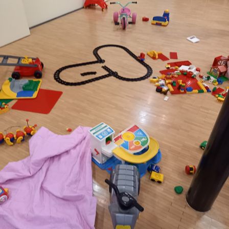 Ein Holzboden mit verschiedenen verstreuten Spielzeugen, darunter ein Zugset, Bausteine, ein Dreirad und andere Spielzeuge. Eine pinke Stoffdecke befindet sich auf der linken Seite.