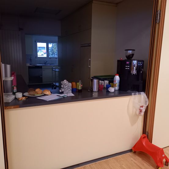 Eine Küchentheke mit einer Kaffeemaschine, einigen Tassen, Flaschen und Lebensmitteln. Ein kleiner roter Spielzeugstuhl steht auf dem Boden.