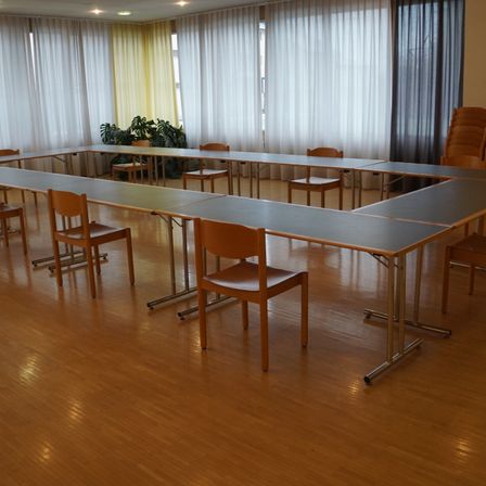 Ein Konferenzraum mit Holzboden und großen Fenstern, die von weißen Vorhängen bedeckt sind. Mehrere lange Tische sind mit Stühlen aufgestellt, und im Hintergrund stehen einige Topfpflanzen.