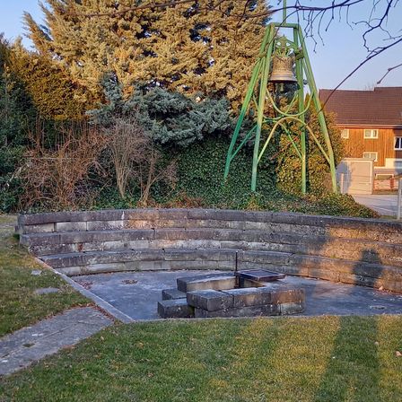 Ein grüner Glockenturm steht in einem Außenbereich, umgeben von einer Steinmauer und Gras. In der Mitte befindet sich ein Steinfeuerplatz. Bäume und ein Gebäude sind im Hintergrund zu sehen.