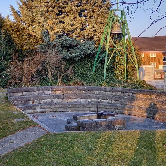 Ein grüner Glockenturm steht in einem Außenbereich, umgeben von einer Steinmauer und Gras. In der Mitte befindet sich ein Steinfeuerplatz. Bäume und ein Gebäude sind im Hintergrund zu sehen.