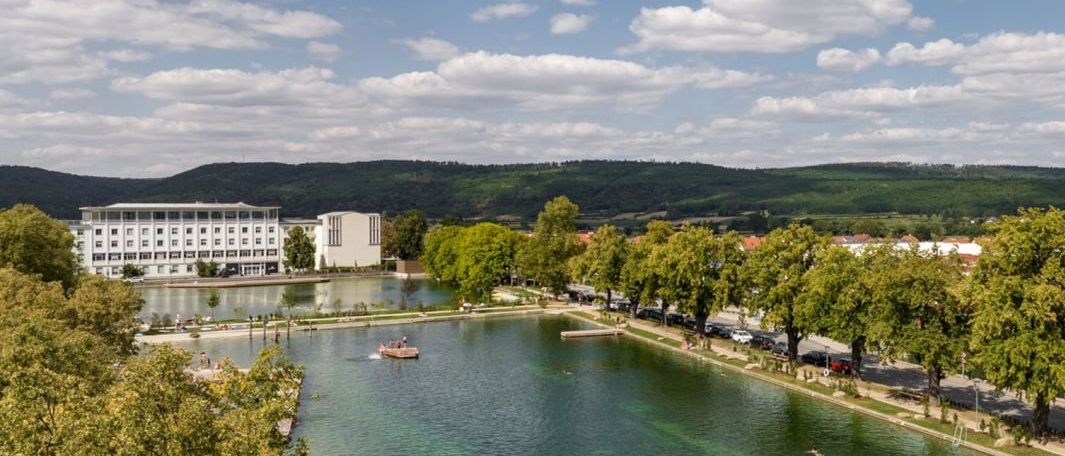 Luftaufnahme eines Sees mit einem schwimmenden Boot, umgeben von Bäumen und Gebäuden, unter einem blauen Himmel mit weißen Wolken.
