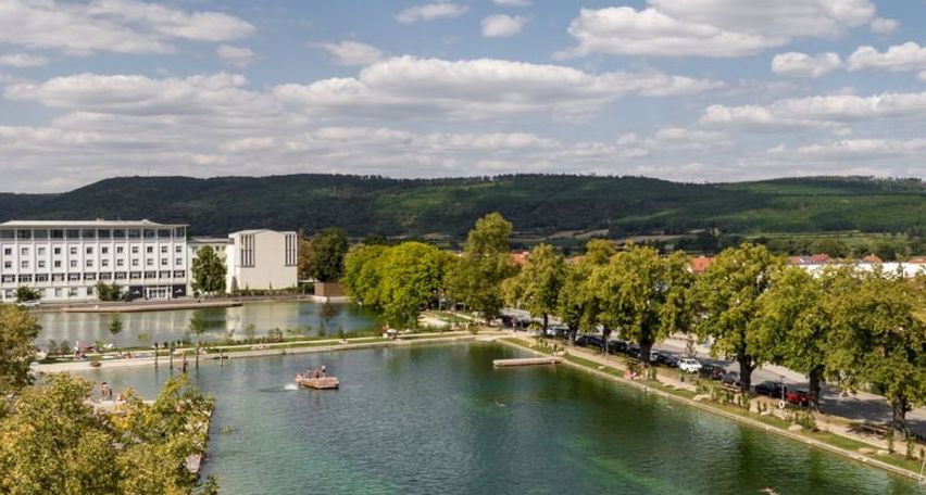 Luftaufnahme eines Sees mit einem schwimmenden Boot, umgeben von Bäumen und Gebäuden, unter einem blauen Himmel mit weißen Wolken.