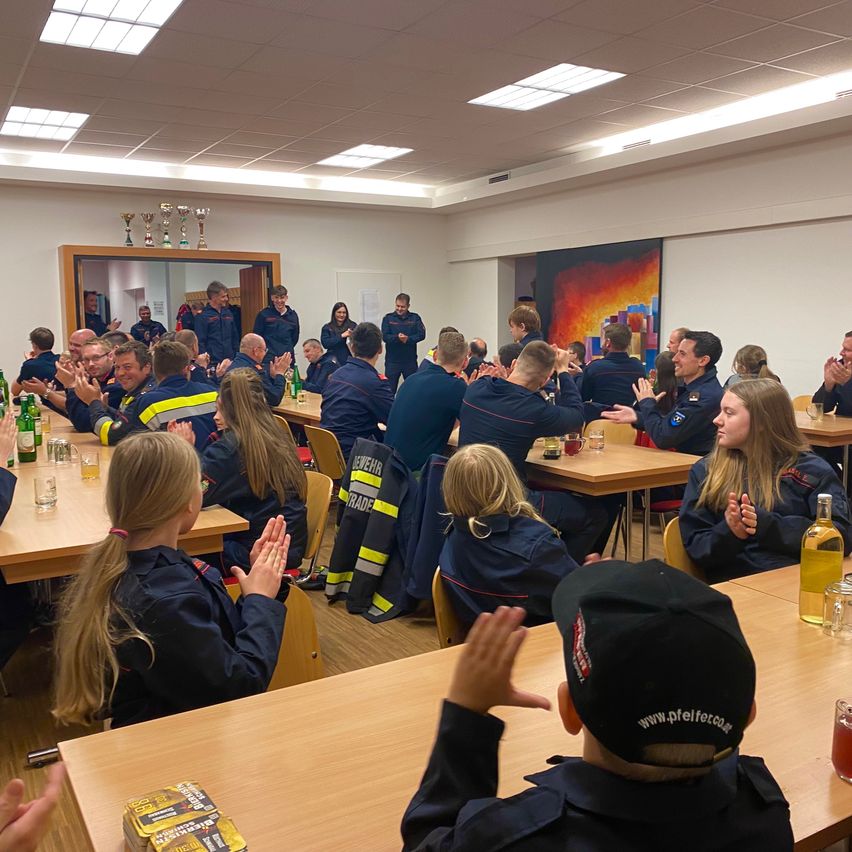 Eine Gruppe von Menschen in blauen Uniformen klatscht in einem Konferenzraum. Tische und Stühle sind aufgestellt. Es gibt Trophäen auf einem Regal und ein großes Gemälde an der Wand. Flaschen, Gläser und eine Schachtel stehen auf den Tischen.