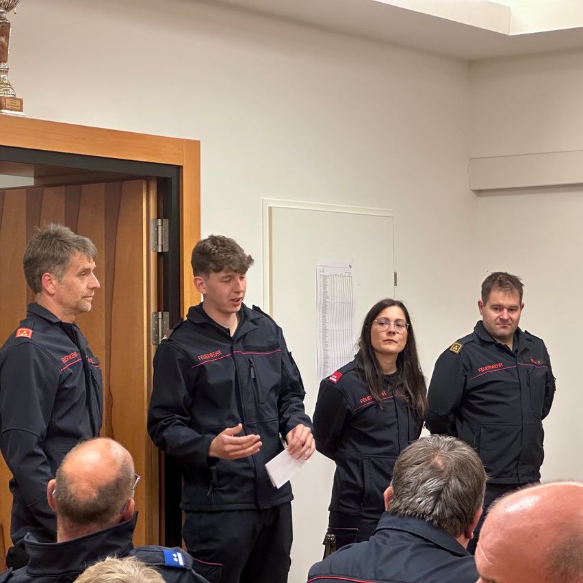 Fünf Personen in Uniformen, wahrscheinlich Feuerwehrleute, stehen in einem Raum. Ein Mann in der Mitte hält ein Stück Papier und spricht zu den anderen. Ein Pokal steht auf einem Regal hinter ihnen.