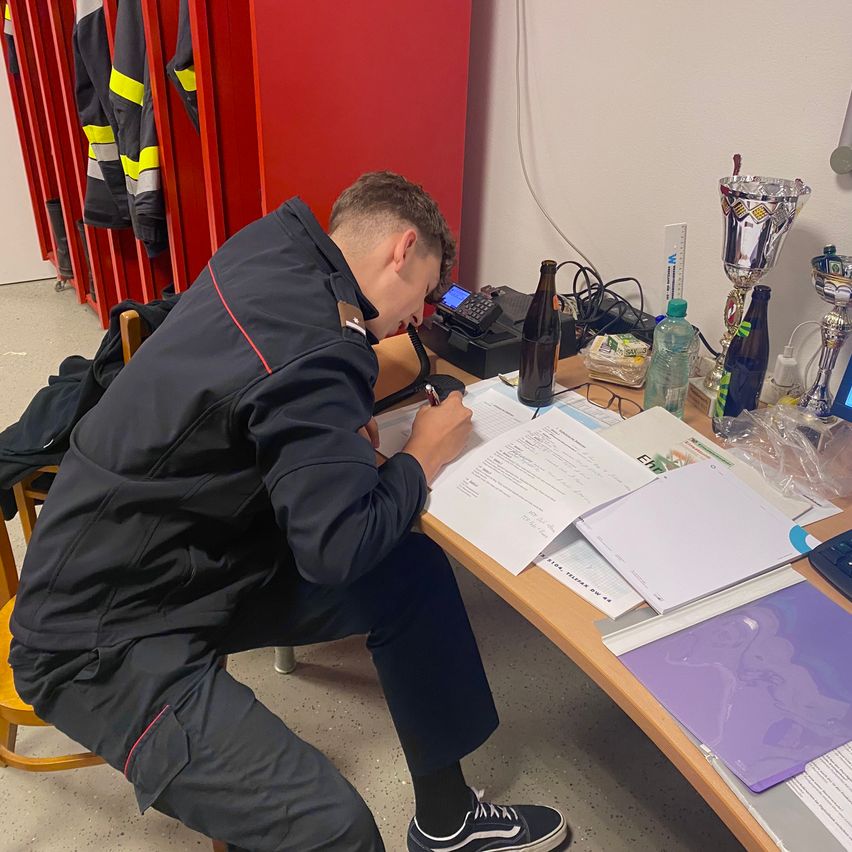 Ein Mann sitzt in einem Raum an einem Schreibtisch und schreibt mit einem Stift auf ein Papier. Er trägt eine Jacke und Hose. Auf dem Schreibtisch befinden sich verschiedene Gegenstände wie ein Pokal, eine Flasche, ein Becher und ein Monitor.