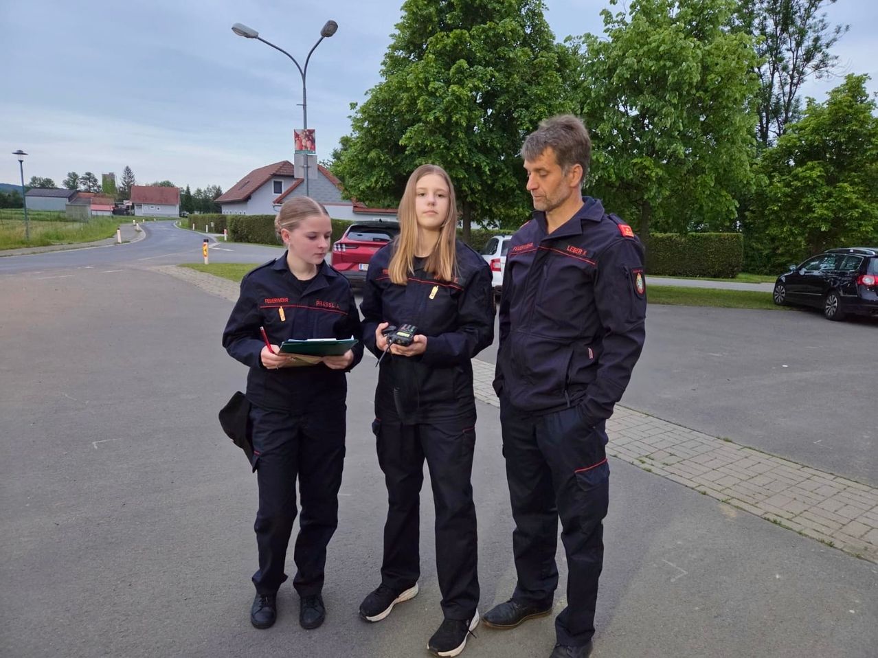 Drei Personen in Uniformen stehen auf einer Straße. Eine Frau hält eine Mappe und einen Stift, und eine andere Frau hält ein Gerät. Ein Mann steht zwischen ihnen. Bäume und Häuser sind im Hintergrund.