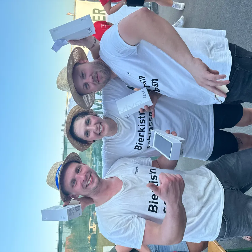Drei Personen in Hüten halten Boxen und tragen weiße T-Shirts mit schwarzem Text, möglicherweise bei einer Veranstaltung. Sie lächeln und posieren für ein Foto.
