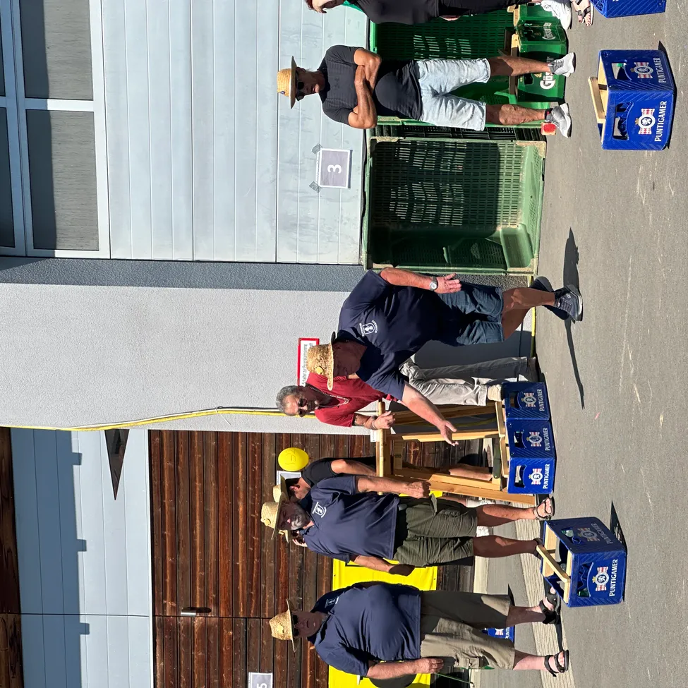 Eine Gruppe von Männern in Hüten und Sonnenbrillen ist in einem Fassklopfwettbewerb engagiert, mit nummerierten Kisten, einer gelben Kugel und einem Gebäude im Hintergrund.