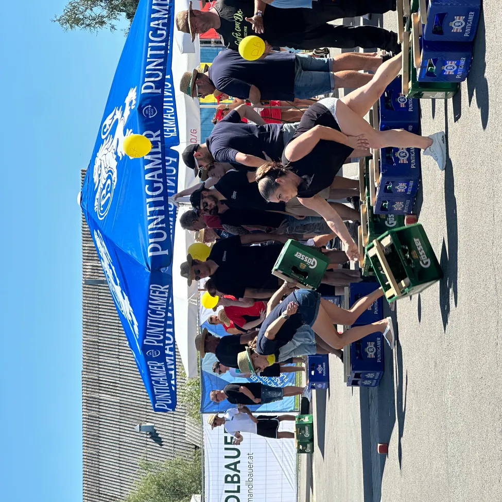 Ein Outdoor-Event mit einem blauen Zelt, auf dem 'Puntigamer' steht. Menschen sind versammelt, einige spielen ein Spiel mit gelben Luftballons, während andere zuschauen. Bierkisten sind in der Nähe gestapelt.