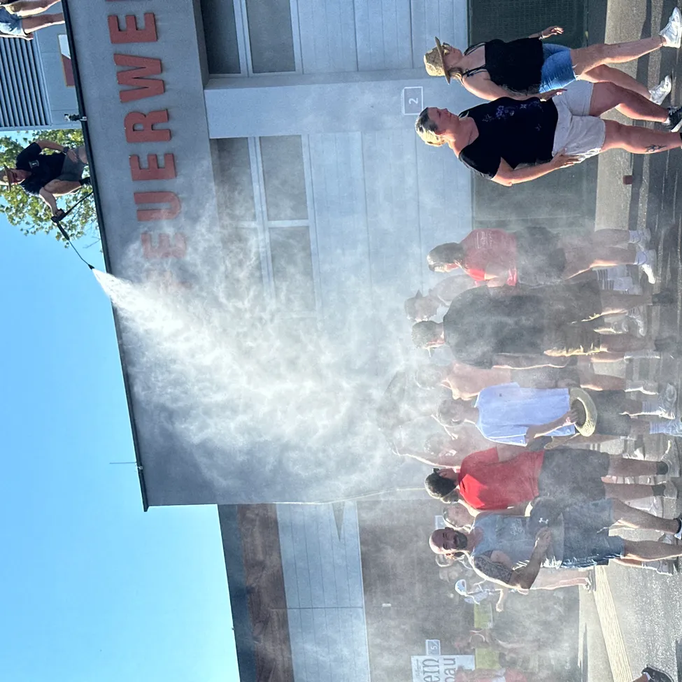 Menschen stehen vor einem Gebäude mit dem Wort Feuerwehr darauf und kühlen sich mit einem Wasserspray ab.