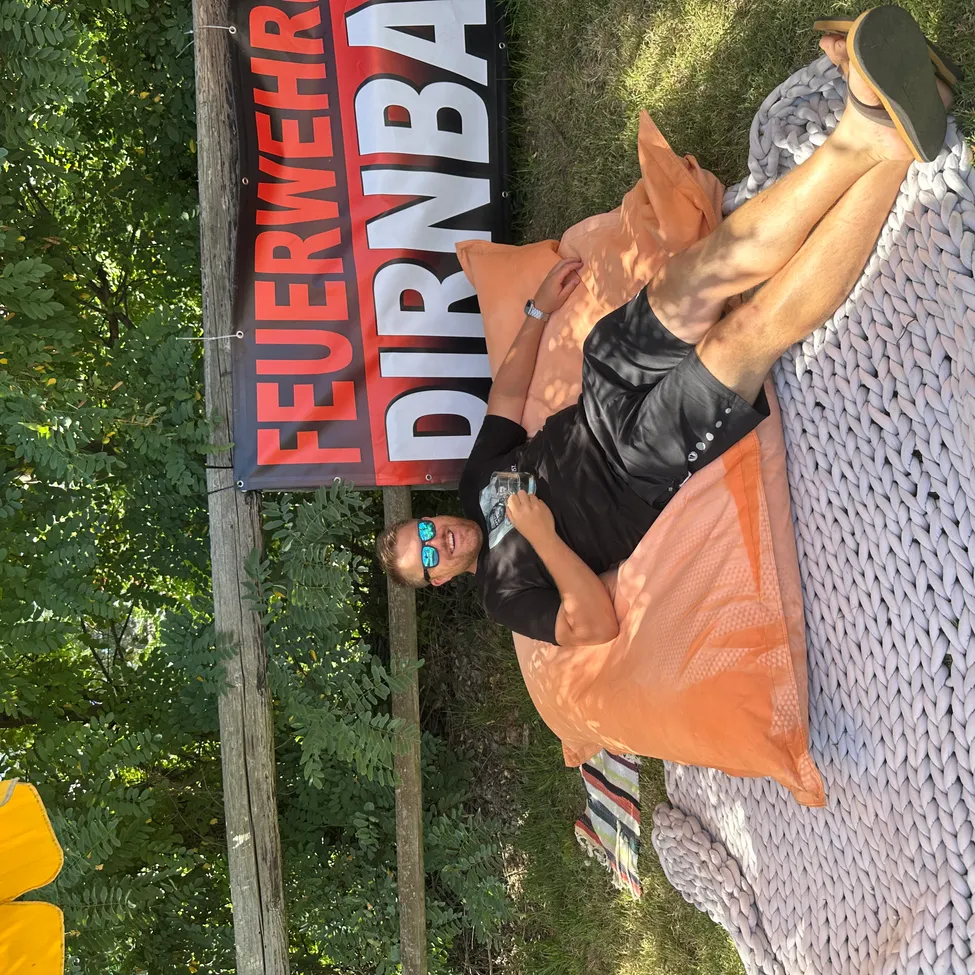 Ein Mann mit einer schwarzen Uhr trägt eine Brille und liegt auf einem orangefarbenen Kissen auf einer Decke. Er stützt seinen Kopf auf seinen Arm.