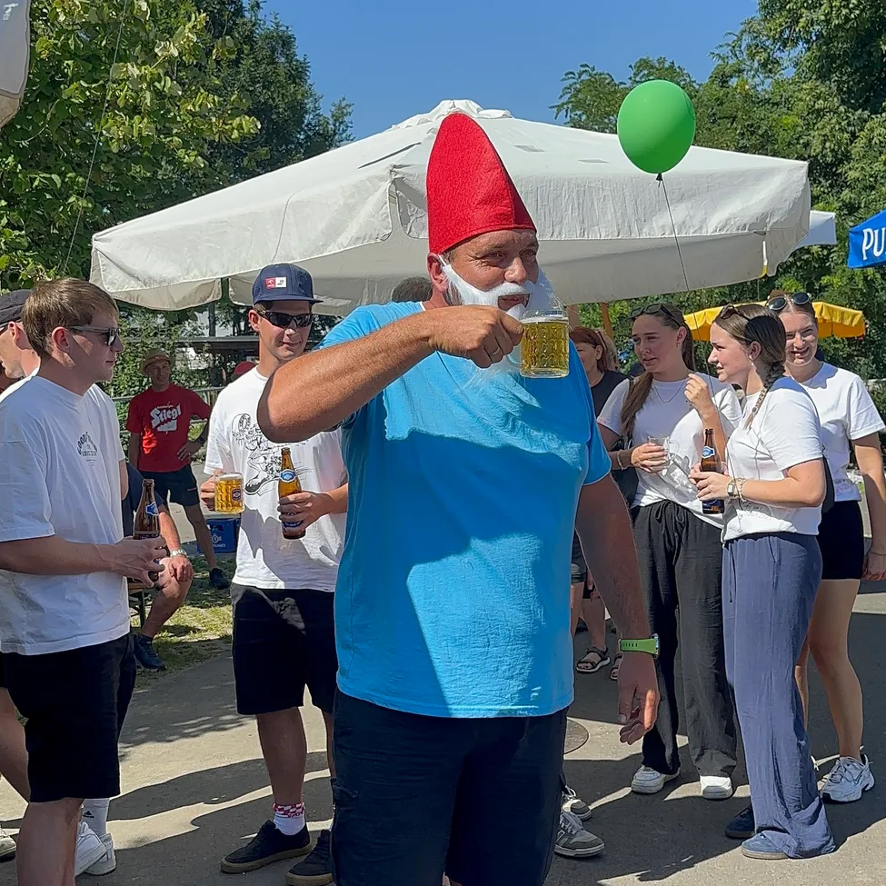 Ein Mann in einem Gnomhut trinkt ein Bier im Park. Mehrere Leute stehen um ihn herum und halten ebenfalls Biere. Sie stehen unter einem großen Zelt.