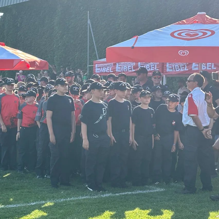 Eine Gruppe von Kindern in schwarzen und roten Uniformen steht auf einem Feld. Ein Mann in einem weißen Hemd steht vor ihnen. Hinter ihnen sind zwei rote und weiße Zelte mit Logos.