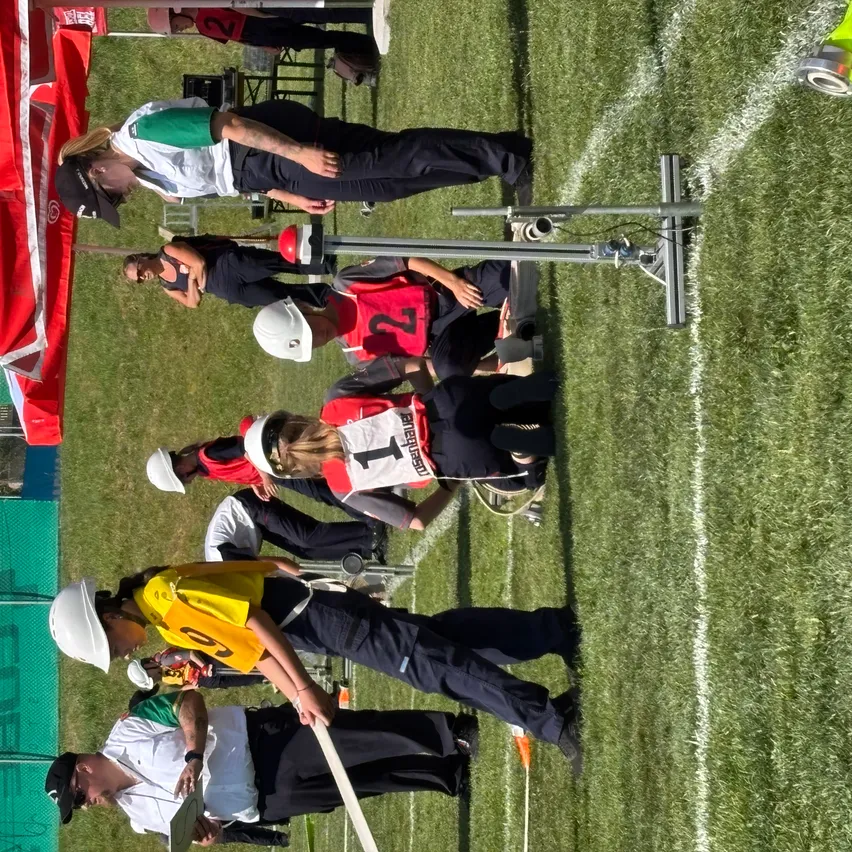 Ein Blick von oben zeigt ein Sportfeld mit mehreren Personen in Uniformen und Helmen. Eine Frau beugt sich über einen Mann, und eine Person in einem weißen Shirt steht in der Nähe. Eine Metallstruktur ist auf dem Feld zu sehen.
