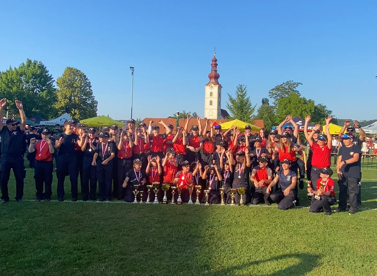 Eine Gruppe von Menschen in roten und schwarzen Uniformen steht auf einem grünen Feld, hält Trophäen und posiert für ein Foto. Dahinter stehen Zelte, ein Turm und Bäume.