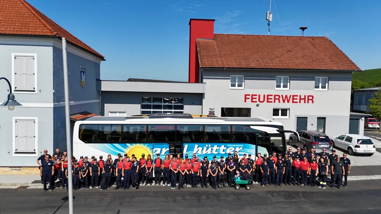Eine Gruppe von Menschen in roten Uniformen posiert vor einem Bus mit einem blau-weißen Design, mit einem Gebäude namens 'Feuerwehr' hinter ihnen.